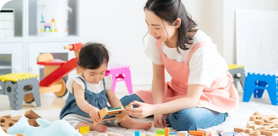 保育士と幼児が積み木のある部屋で遊ぶ様子